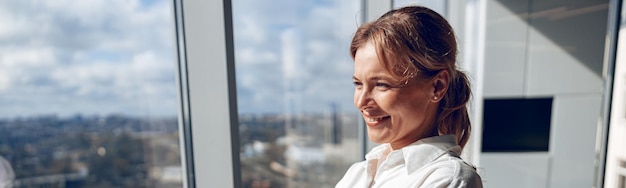 Foto empresária madura sorridente em pé perto da grande janela no escritório corporativo moderno