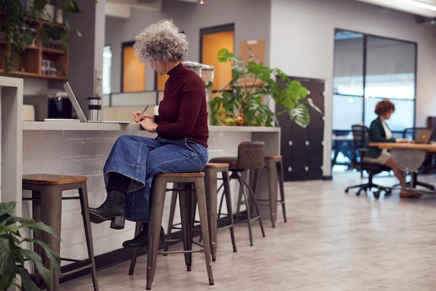 Empresaria madura con taza de viaje trabajando en la computadora portátil en la oficina moderna a través del teléfono móvil