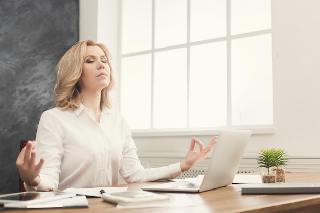 Empresária meditando no escritório. Retrato de mulher de meia-idade com roupa formal se acalmando no local de trabalho, copie o espaço