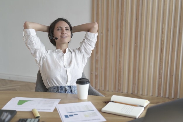 Empresaria meditativa en auriculares sentada en posición relajada con las manos detrás de la cabeza