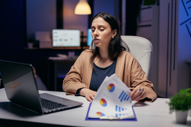 Empresaria mirando en el monitor de la computadora portátil tratando de encontrar soluciones. Mujer revisando notas en el portapapeles mientras trabajaba en un proyecto importante haciendo horas extraordinarias.