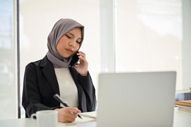 Empresária muçulmana asiática com lenço na cabeça falando ao telefone com seu parceiro de negócios