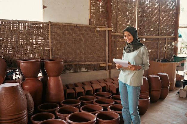 Empresária muçulmana asiática sorridente segurando a prancheta enquanto na barraca de cerâmica