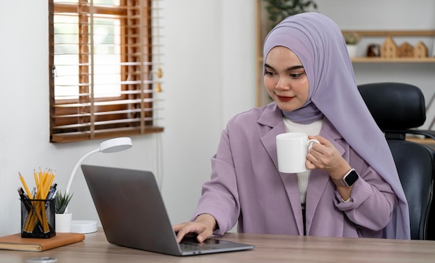 Empresária muçulmana sorridente segurando a xícara de café sentada à mesa no escritório em casa Jovem aluna islâmica usando hijab e roupa casual inteligente usando laptop no escritório