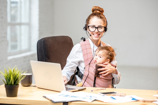 Foto empresaria multitarea que trabaja con auriculares y computadora portátil sentada con su hijo en el interior de la oficina blanca