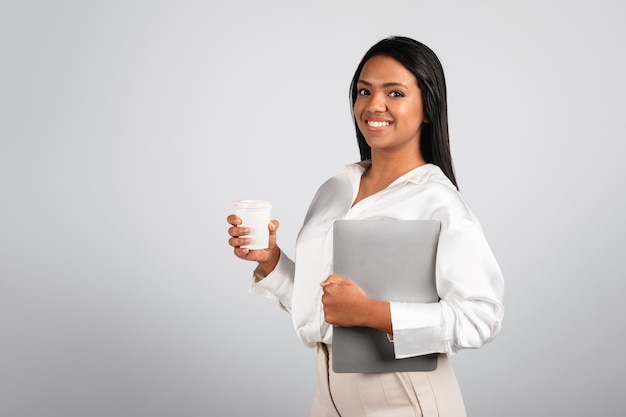 Empresária negra milenar sorridente e confiante em blusa branca com laptop e xícara de café para viagem