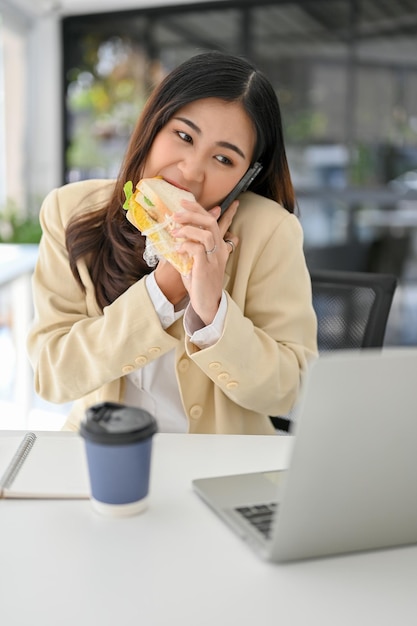 Empresária ocupada comendo um sanduíche enquanto olha para a tela do laptop e fala ao telefone