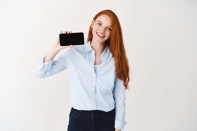 Empresaria pelirroja alegre mostrando la pantalla del teléfono inteligente, mostrando la pantalla del teléfono en blanco y sonriendo, de pie sobre la pared blanca