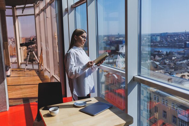 Empresária pensativa com o bloco de notas vestido com roupa formal em pé perto da janela panorâmica na empresa corporativa e pensando em investimento comercial empresária pensativa em óculos