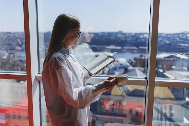 Empresária pensativa com o bloco de notas vestido com roupa formal em pé perto da janela panorâmica na empresa corporativa e pensando em investimento comercial empresária pensativa em óculos