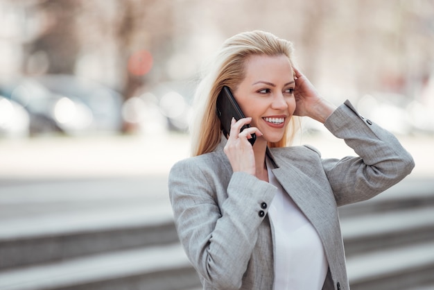 Empresaria positiva en el desgaste formal que habla en el teléfono en la ciudad en la primavera.