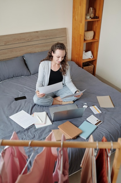 Empresaria positiva sentada en la cama en casa y trabajando con documentos, vista desde arriba