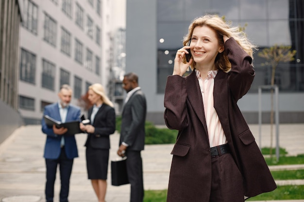 Empresaria rubia de pie y hablando por teléfono frente a la oficina moderna