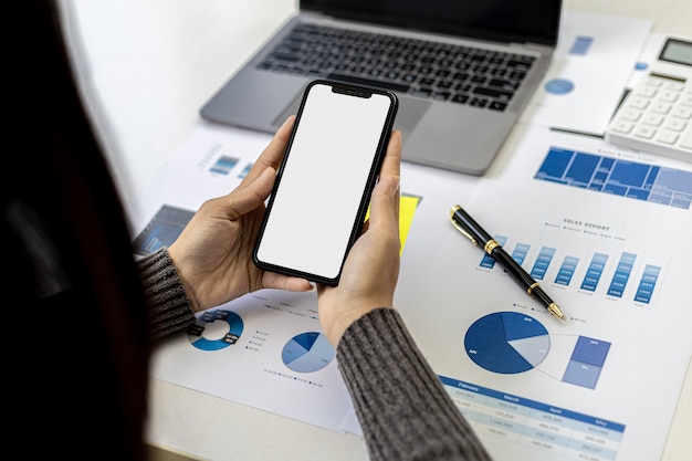 Empresária segurando smartphone na mesa com documentos e laptop colocado, ela está olhando dados do celular, tela em branco do celular para gráficos de publicidade. O conceito de uso de tecnologia