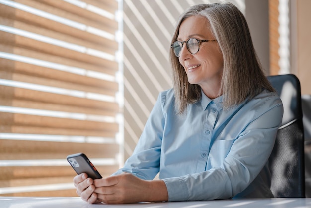 Empresária sênior sorridente segurando um celular lendo mensagens de texto on-line