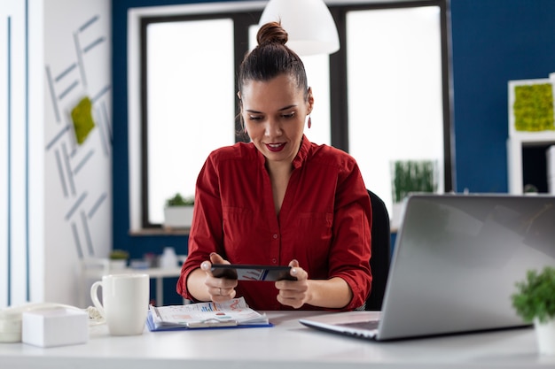 Empresária, sentada à mesa no escritório corporativo, jogando videogame usando smartphone empresário t ...