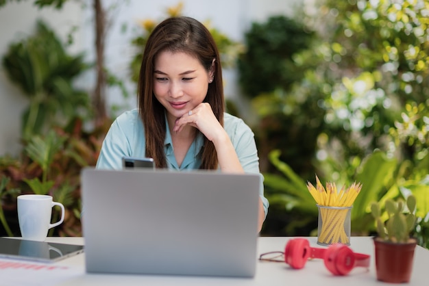 La empresaria sentada en el escritorio de trabajo con el teléfono inteligente y la computadora portátil se conectan a internet en la esquina del fondo verde del pequeño jardín en casa. Concepto de nueva gente normal y trabajo autónomo en casa.