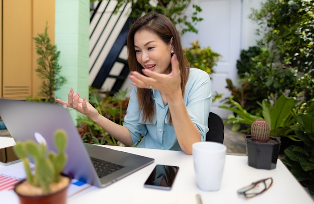 La empresaria sentada en el pequeño jardín de la casa de la esquina en el escritorio de trabajo usando la computadora portátil se conecta a la reunión en línea y levanta la mano para explicar a los asistentes. Concepto de nueva gente normal y trabajo en casa.