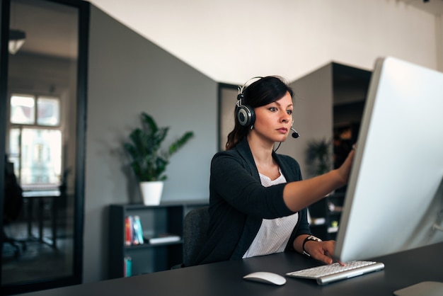 Empresaria seria con las auriculares que solucionan problemas mientras que trabaja en la computadora en oficina moderna.