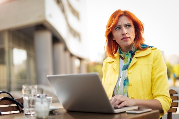 Empresária séria trabalhando no laptop em uma pausa para o café em um café de rua. Olhando para longe.