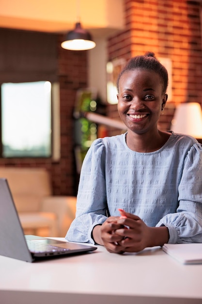 Empresaria sonriente que trabaja en la computadora portátil en el lugar de trabajo en la oficina en casa. Joven gerente de proyecto de equipo remoto afroamericano en el lugar de trabajo en una habitación contemporánea con grandes ventanas