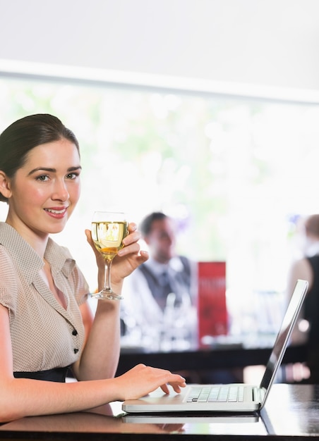 Empresaria sonriente que usa la computadora portátil mientras que sostiene el vidrio de vino