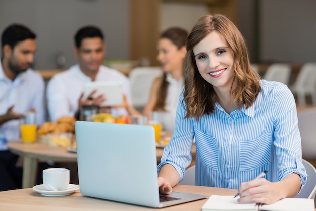 Empresaria sonriente trabajando en equipo portátil mientras toma un café en la cafetería de la oficina