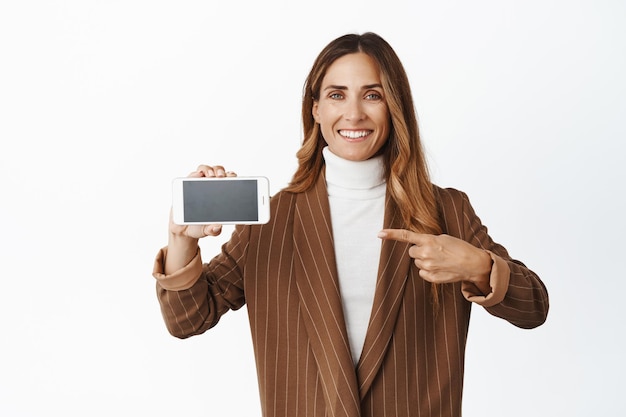 Empresária sorridente apontando o dedo para a tela horizontal do smartphone mostrando o aplicativo em pé sobre fundo branco