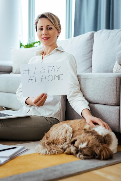 Empresária sorridente com um cachorro segurando um cartaz com a hashtag eu fico em casa enquanto trabalhava em casa