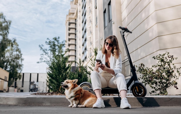 Empresária sorridente de terno branco sentado na scooter elétrica e usando o telefone durante a caminhada com cachorro Welsh Corgi Pembroke na cidade
