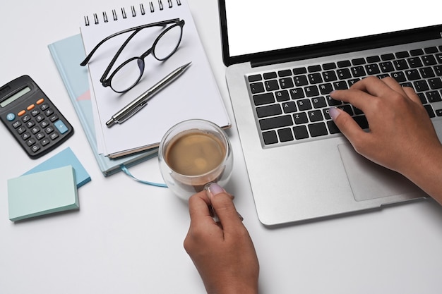 Empresaria sosteniendo la taza de café y trabajando con ordenador portátil en el escritorio de oficina blanco.