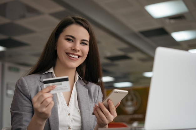 Empresaria con tarjeta de crédito, utilizando el teléfono inteligente para compras en línea, sentado en la oficina