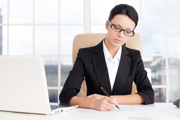 Empresaria en el trabajo. Confianza joven empresaria escribiendo algo en papel mientras está sentada en su lugar de trabajo