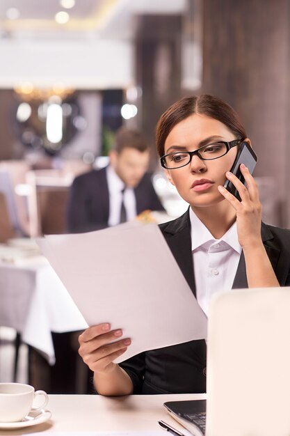 Empresaria en el trabajo. Mujer joven pensativa en ropa formal hablando por teléfono y mirando los documentos mientras está sentado en el restaurante