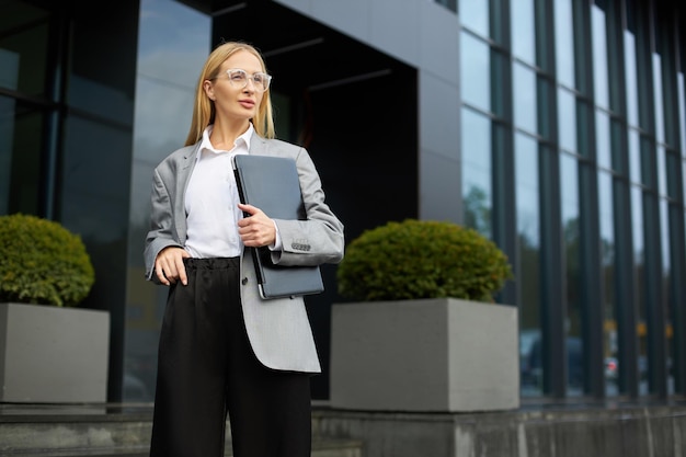 Empresária usando óculos elegantes segurando laptop olhando para longe em pé na rua