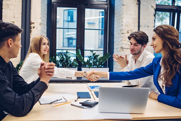 Empresarias exitosas dándose la mano mientras los empresarios aplaudiendo en la mesa en la oficina