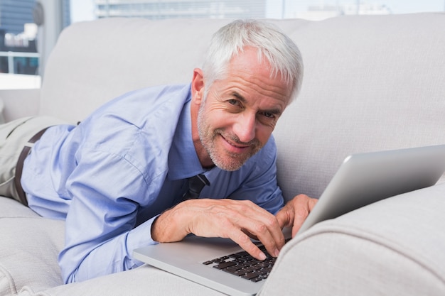 Empresario acostado en el sofá usando la computadora portátil y sonriendo a la cámara