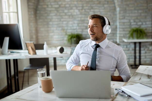 Empresário adulto médio assinando enquanto ouve música em fones de ouvido e trabalhando no laptop no escritório