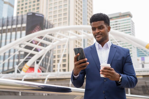 Empresario africano al aire libre mediante teléfono móvil y sosteniendo la taza de café para llevar, tiro horizontal