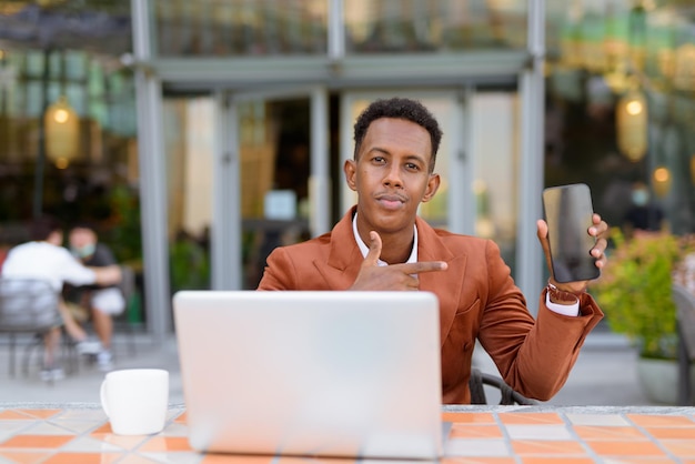 Empresário africano ao ar livre em um café usando um laptop e mostrando o celular com espaço de cópia