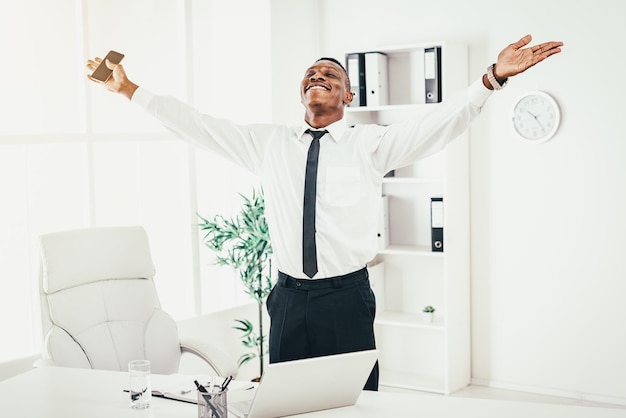 Empresário africano bem sucedido comemorando o sucesso com os braços levantados no escritório moderno.