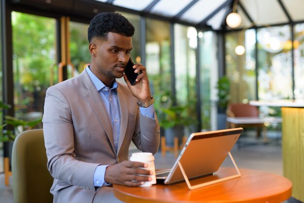 Empresario africano en la cafetería con ordenador portátil mientras habla por teléfono