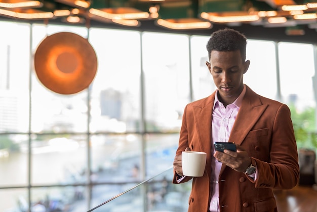 Empresario africano en cafetería mediante teléfono móvil y sosteniendo la taza de café