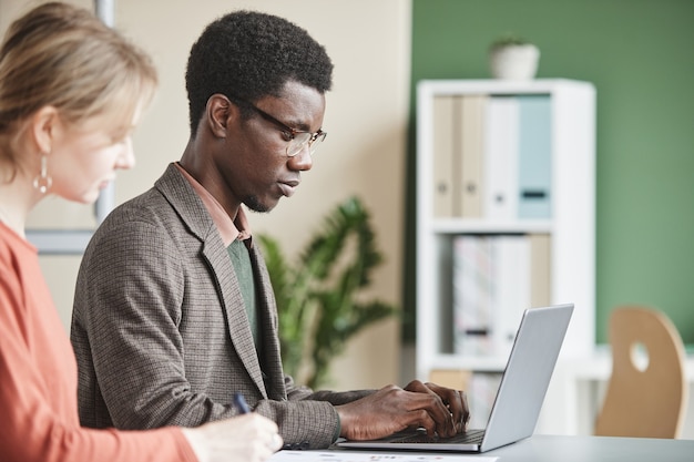 Empresário africano digitando no laptop com seu colega sentado perto dele à mesa no escritório