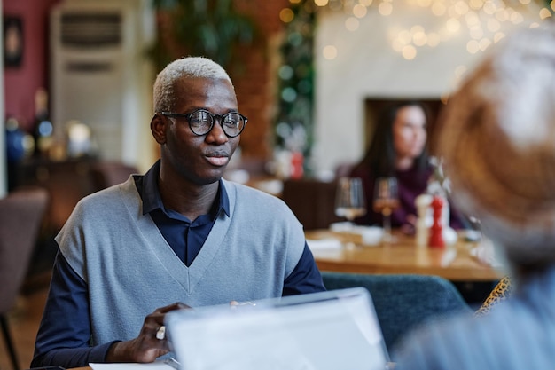 Empresário africano em óculos, sentado à mesa de um café, tendo uma reunião de negócios com seus colegas