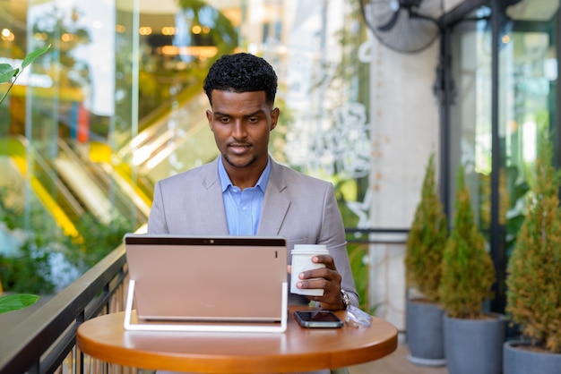 Empresário africano em um café ao ar livre, usando um laptop e segurando uma xícara de café