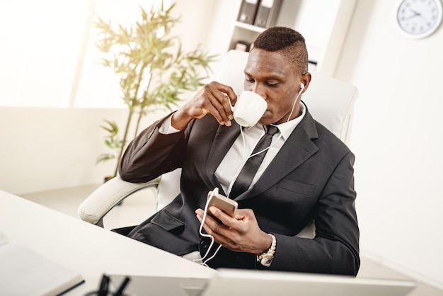 Empresário africano está sentado no escritório, tomando café e ouvindo música em fones de ouvido.