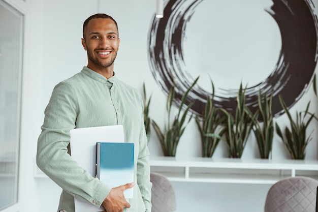 Empresário africano sorridente em pé no escritório moderno e segurando laptop