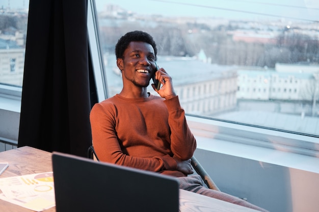 Empresário africano sorridente falando no celular enquanto está sentado à mesa em frente ao laptop no escritório