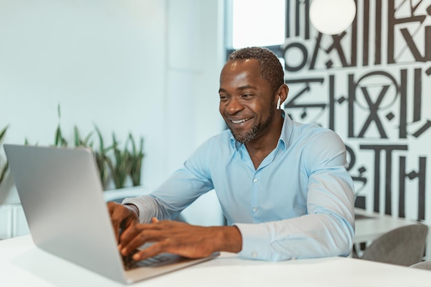 Empresário africano sorridente trabalhando no laptop enquanto está sentado no escritório moderno